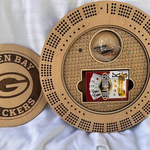 May include: A wooden cribbage board with a circular design, featuring a storage compartment with a deck of cards labeled "FULL HOUSE". The board has a lid with the Green Bay Packers logo.