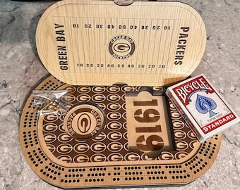 May include: A wooden Green Bay Packers cribbage board with a football field design on the lid, a 1919 cutout, a team logo, a small plastic bag containing metal pegs, and a standard deck of Bicycle playing cards resting on the game board surface.