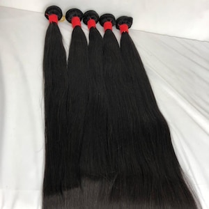 Puede incluir: Cinco paquetes de extensiones de cabello largo, liso y negro azabache. Cada paquete está asegurado en la parte superior con una banda roja vibrante y gruesa, manteniendo los mechones alineados y cayendo con una textura suave y uniforme.
