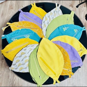 May include: A collection of 10 colorful fabric pouches with zipper closures. The pouches are arranged in a circle on a black surface. The pouches are made of different fabrics and have various patterns and colors, including yellow, green, blue, purple, and white.