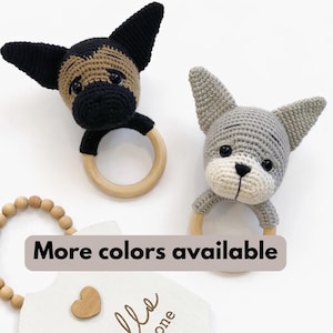 May include: Two crochet dog-shaped baby rattles. One is black and tan, the other is gray and white. Each has a wooden ring for the baby to grasp. The text "More colors available" is displayed on a banner.