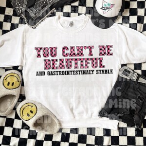 May include: White sweatshirt with pink and black checkered text that reads "You can't be beautiful and gastrointestinally stable."