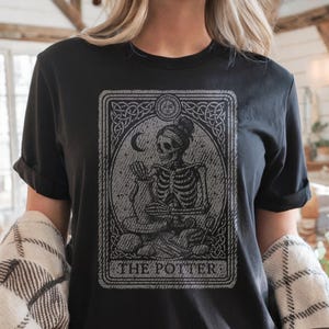 May include: Black t-shirt featuring a tarot card design with a skeleton figure. The design is in shades of gray and includes the text "THE POTTER" at the bottom. The shirt has short sleeves and a crew neck.