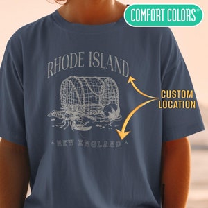 May include: A blue t-shirt with a light grey print of a lobster trap and the text "RHODE ISLAND" and "NEW ENGLAND". The text "CUSTOM LOCATION" is also printed on the shirt.