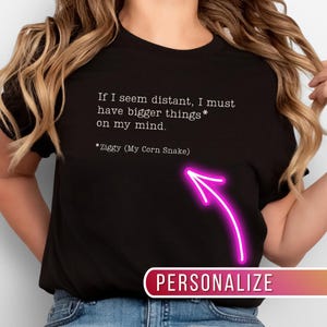 May include: Black t-shirt with white text: "If I seem distant, I must have bigger things* on my mind. *Ziggy (My Corn Snake)". A pink arrow points to the word "PERSONALIZE". The word "Black" is at the bottom.