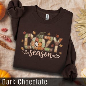 May include: Dark chocolate brown sweatshirt with the words "Cozy season" and a smiling pumpkin graphic. The design features fall leaves and a patchwork pumpkin. A cosy autumn-themed item of clothing.