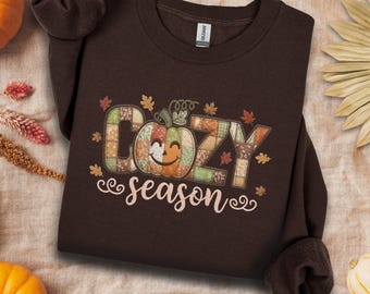 May include: Dark chocolate brown sweatshirt with the words "Cozy season" and a smiling pumpkin graphic. The design features fall leaves and a patchwork pumpkin. A cosy autumn-themed item of clothing.