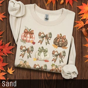 May include: A sand-colored sweatshirt featuring a fall-themed design. The design includes various pumpkins with plaid and floral patterns, adorned with bows. The word "Sand" is at the bottom of the image. Autumn leaves and acorns are scattered around the sweatshirt.