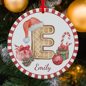May include: Round Christmas ornament with a red and white border. The ornament features a gingerbread-style letter "E" wearing a Santa hat, a candy cane, and gift boxes. The name "Emily" is written in script.