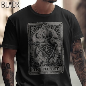 Puede incluir: Una camiseta negra con un estampado gráfico de un esqueleto tocando la flauta dentro de un marco rectangular. El diseño incluye un sol, una luna creciente y un murciélago. El texto "THE FLUTIST" está impreso en la parte inferior del dibujo.