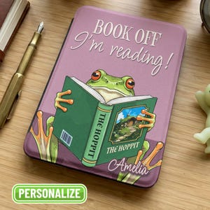 May include: A personalized tablet case with a frog reading "The Hoppit." The case is lavender with the text "Book off I'm reading!" and the name "Amelia." The frog is green with orange eyes and is holding a green book.