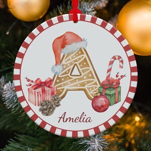 May include: A circular Christmas decoration with a red and white striped edge. It displays a gingerbread-style letter "A" wearing a Santa hat, a candy cane, gifts, and the name "Amelia".