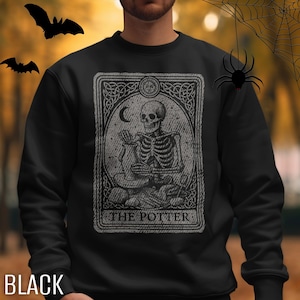 May include: Black sweatshirt featuring a tarot card design with a skeleton illustration. The card reads "THE POTTER" and includes a crescent moon and ornate border. Bats and a spider web are in the background. The word "BLACK" is at the bottom.