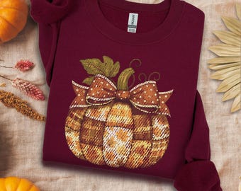 May include: Maroon sweatshirt with a pumpkin graphic. The pumpkin has a plaid pattern and a large bow with green leaves. The word "Maroon" is printed at the bottom of the image. The sweatshirt is folded and displayed on a neutral surface.