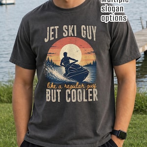 Puede incluir: Una camiseta gris carbón de manga corta con un gráfico de una persona en una moto de agua frente a una puesta de sol. El texto dice JET SKI GUY like a regular guy BUT COOLER. La camiseta tiene una etiqueta Comfort Colors y el color Pepper.