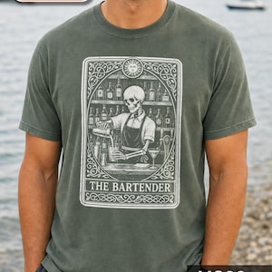 May include: A charcoal grey short-sleeved t-shirt featuring a graphic print of a skeleton bartender in an apron pouring a drink. The design is framed like a tarot card with the text THE BARTENDER at the bottom and a sun icon at the top.