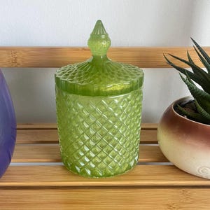 May include: A green glass candle jar with a diamond pattern and a pointed lid. The jar is sitting on a wooden surface.