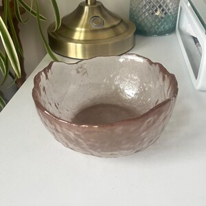 Könnte beinhalten: Eine kleine, klare Glasschüssel mit einer strukturierten, mattierten Oberfläche. Die Schüssel hat einen leicht rosafarbenen Farbton und einen leicht gewellten Rand.