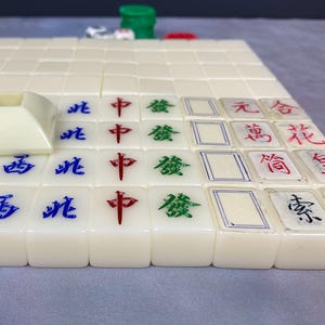 May include: A close-up of a Mahjong game in progress. The ivory-colored tiles are arranged on a surface, displaying Chinese characters in red, green, and blue. A white tile holder is visible in the foreground, with other game pieces in the background.