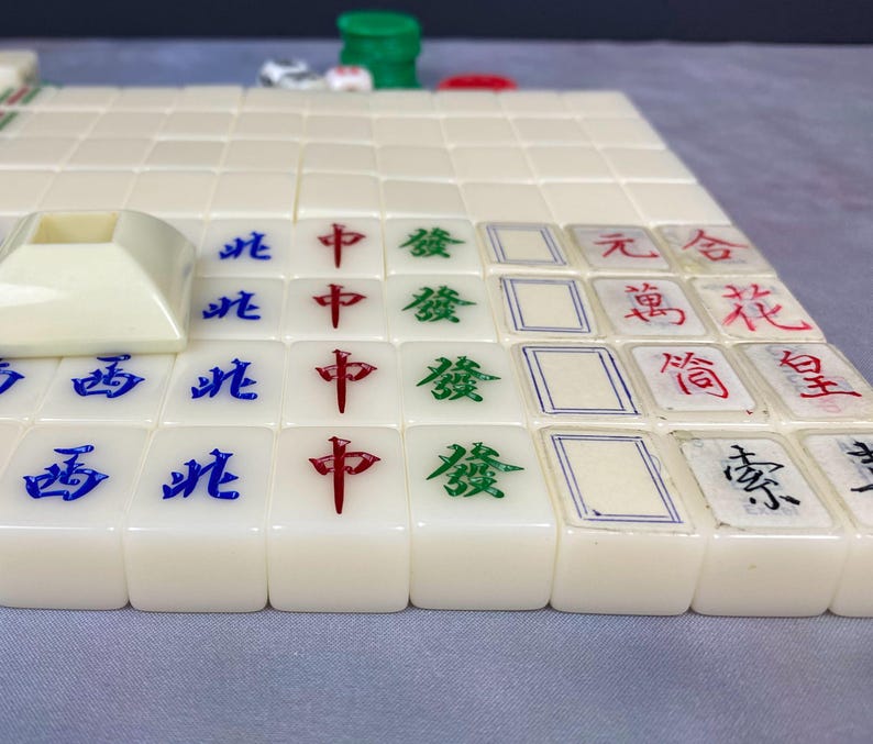 May include: A close-up of a Mahjong game in progress. The ivory-colored tiles are arranged on a surface, displaying Chinese characters in red, green, and blue. A white tile holder is visible in the foreground, with other game pieces in the background.