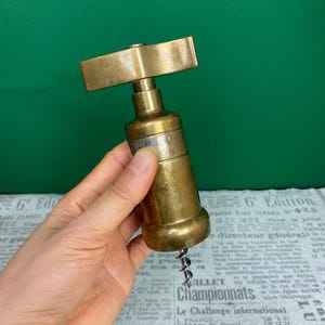 May include: A vintage brass corkscrew with a rectangular handle and a cylindrical body. The corkscrew features a spiral worm for extracting corks. The metal has a warm, aged patina, suggesting it is an antique. The design is simple and functional.