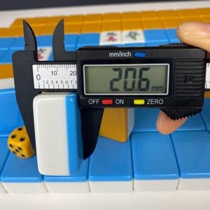 Puede incluir: Una ficha de Mahjong blanca y azul se mide con un calibrador digital. La ficha mide 2,06 cm de ancho. Un dado amarillo con puntos negros y otras fichas de Mahjong est&aacute;n en el fondo.