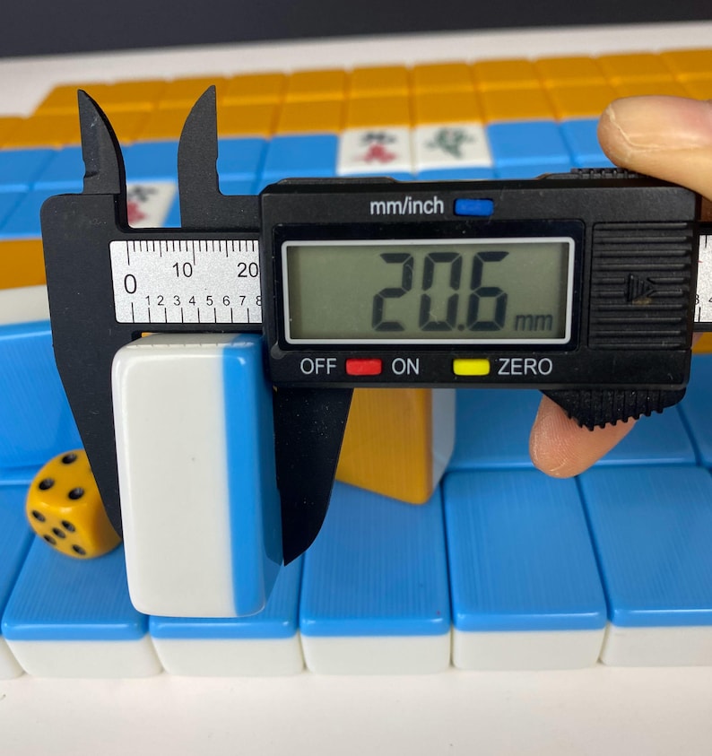 Puede incluir: Una ficha de Mahjong blanca y azul se mide con un calibrador digital. La ficha mide 2,06 cm de ancho. Un dado amarillo con puntos negros y otras fichas de Mahjong est&aacute;n en el fondo.
