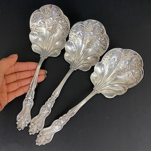 Puede incluir: Tres cucharas de servir plateadas ornamentadas con un diseño floral y de uvas. Las cucharas tienen un cuenco grande en forma de hoja y un mango detallado. Las cucharas se exhiben sobre una superficie negra.