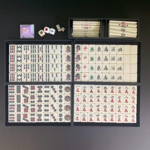 May include: A complete Mahjong set with ivory-colored tiles, dice, and a wind indicator. The tiles are arranged in black trays, displaying various symbols and characters in red and black. The set includes dice and a small, square wind indicator.