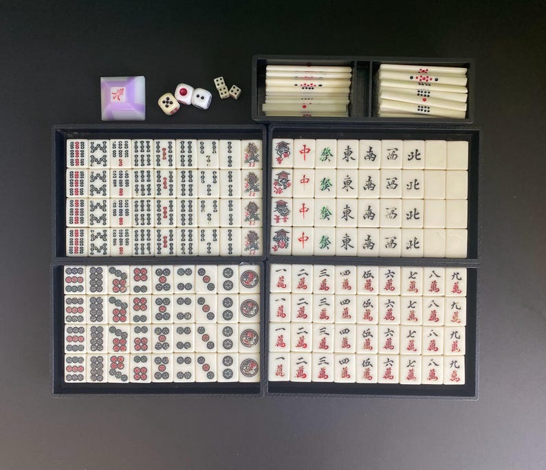 May include: A complete Mahjong set with ivory-colored tiles, dice, and a wind indicator. The tiles are arranged in black trays, displaying various symbols and characters in red and black. The set includes dice and a small, square wind indicator.