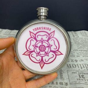 May include: A silver-coloured hip flask with a white face displaying a pink rose emblem and the word "YORKSHIRE". The flask has a silver cap and is held in a hand.