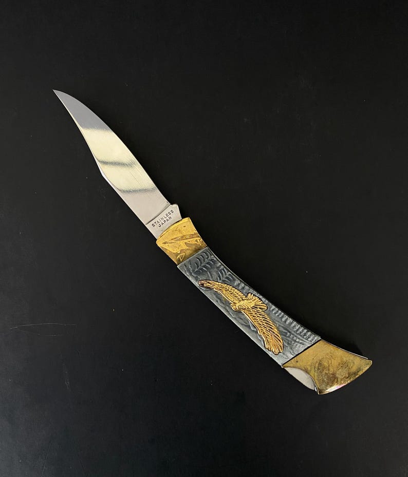May include: A stainless steel folding knife with a silver blade and a handle featuring a gold eagle design. The handle has gold and silver accents. The blade is marked "STAINLESS JAPAN".