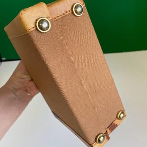 May include: A tan canvas box-shaped bag with leather trim and brass-colored metal studs. The bag has a rectangular shape and is held at an angle, showcasing its construction and materials. The bag's corners are reinforced with leather.