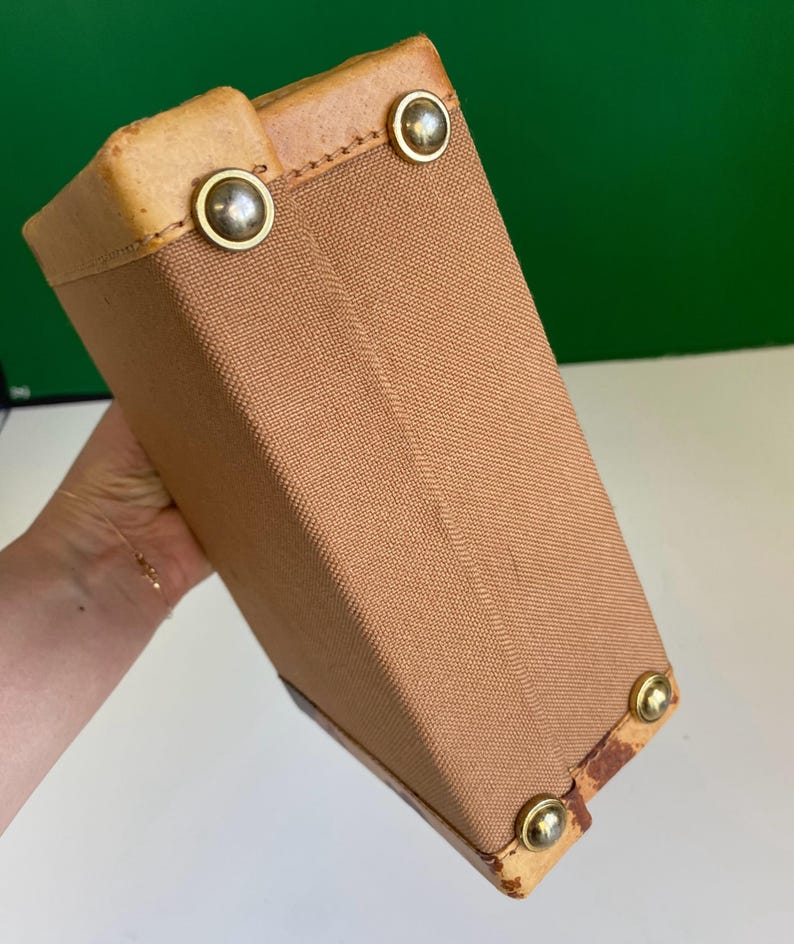 May include: A tan canvas box-shaped bag with leather trim and brass-colored metal studs. The bag has a rectangular shape and is held at an angle, showcasing its construction and materials. The bag's corners are reinforced with leather.