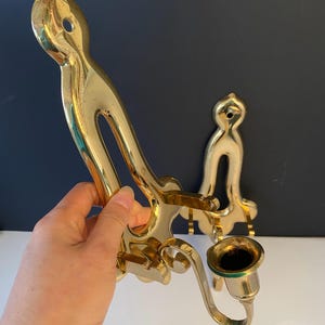 May include: A pair of vintage brass wall sconces. The sconces have a curved design with a candle holder. The brass has a polished finish. One sconce is held by a hand.
