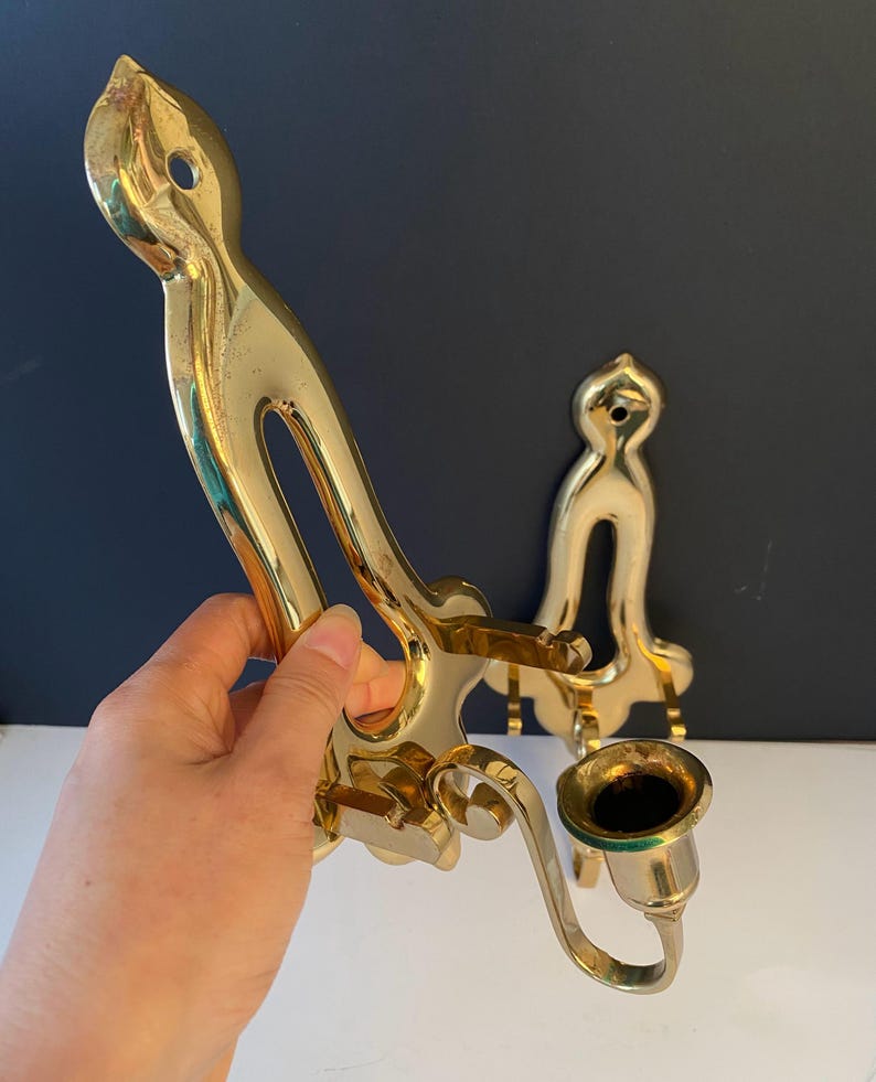 May include: A pair of vintage brass wall sconces. The sconces have a curved design with a candle holder. The brass has a polished finish. One sconce is held by a hand.