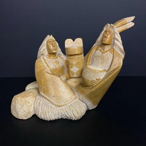 May include: A tan-colored sculpture depicting two figures in traditional Native American attire. The figures are seated, with one holding a vessel and the other a decorative object. The sculpture has detailed carvings and a matte finish.
