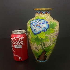 May include: A cloisonné vase featuring a cream-coloured background with a fine wire pattern, decorated with large blue and white peony flowers and green leaves. The vase has a gold-toned rim and is positioned next to a 355 ml red soda can.
