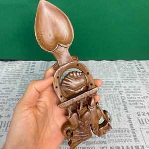 Könnte beinhalten: Ein handgeschnitzter Holzlöffel mit herzförmiger Schüssel und aufwendigen Schnitzereien. Der Löffel hat ein Hufeisen-Design und eine Figur mit fledermausartigen Flügeln. Das Holz hat einen warmen, braunen Farbton.