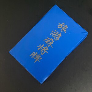 Puede incluir: Una caja rectangular azul brillante con caracteres japoneses dorados. La caja est&aacute; ligeramente inclinada, revelando su superficie superior y bordes. El texto parece ser un t&iacute;tulo o una descripci&oacute;n.