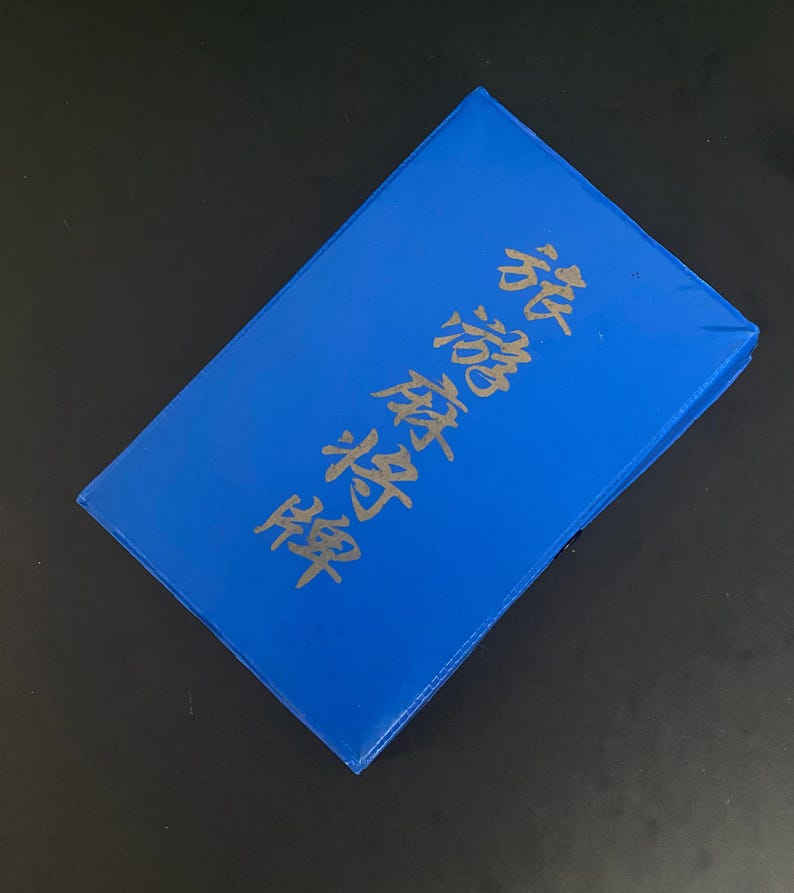 Puede incluir: Una caja rectangular azul brillante con caracteres japoneses dorados. La caja est&aacute; ligeramente inclinada, revelando su superficie superior y bordes. El texto parece ser un t&iacute;tulo o una descripci&oacute;n.