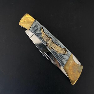 May include: A folding pocket knife with a silver blade and a handle featuring a gold eagle emblem. The handle has a gray background with a decorative pattern. The ends of the handle are gold-colored.