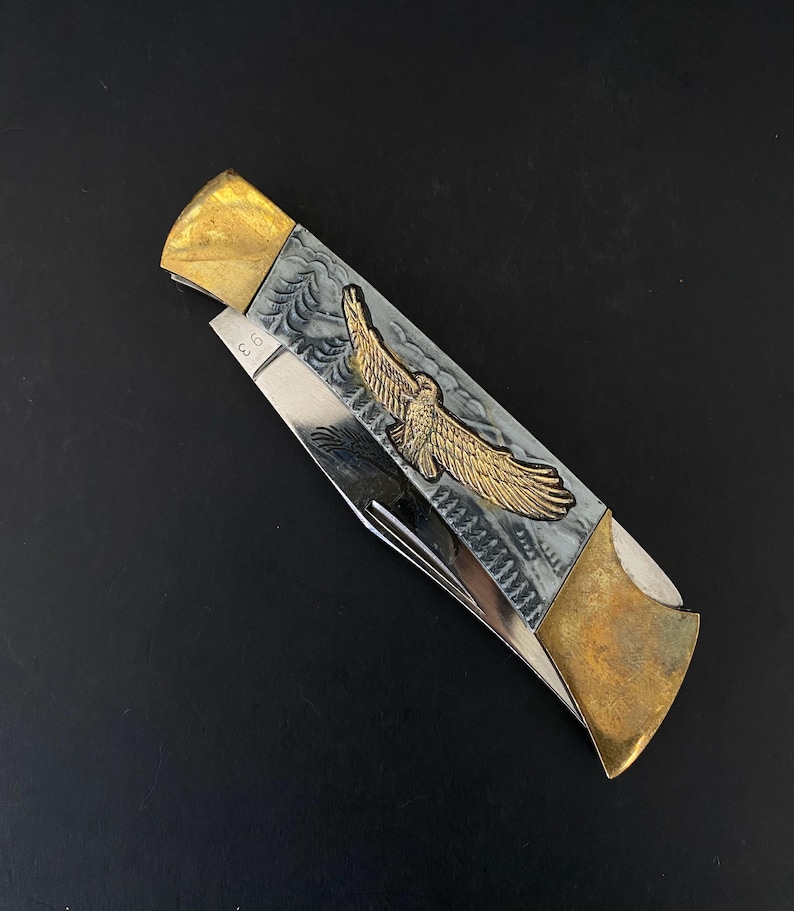 May include: A folding pocket knife with a silver blade and a handle featuring a gold eagle emblem. The handle has a gray background with a decorative pattern. The ends of the handle are gold-colored.