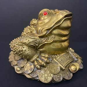 May include: A gold-coloured toad figurine with a textured, bumpy back and a single bright red eye. The toad is perched atop a base composed of numerous round coins and a scroll, each featuring embossed Chinese characters and symbols.