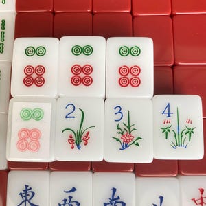 May include: A close-up of a Mahjong set with white tiles featuring red and green circular designs, and floral and number illustrations. Red tiles form the background.