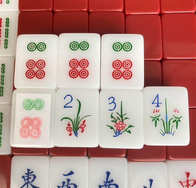 May include: A close-up of a Mahjong set with white tiles featuring red and green circular designs, and floral and number illustrations. Red tiles form the background.
