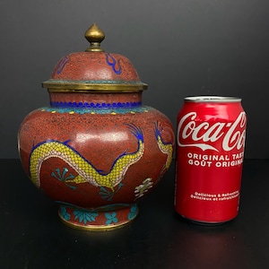 May include: A red and gold decorative jar with a lid, featuring a dragon design. The jar has a brass-coloured rim and a gold finial on the lid. The jar is next to a Coca-Cola can.