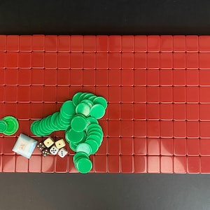 May include: A red rectangular game board with a grid of red tiles. Green circular game pieces and dice are scattered across the board. A small white pouch is also present. The game pieces are in a variety of shapes and sizes.
