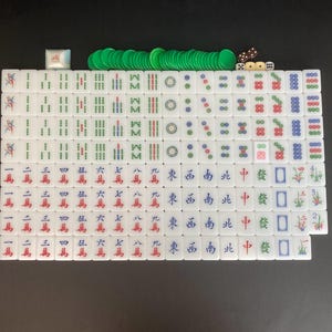 May include: A complete Mahjong set with white tiles featuring Chinese characters and symbols in red, green, and blue. Includes green chips, dice, and a white wind indicator. The set is arranged on a black surface.