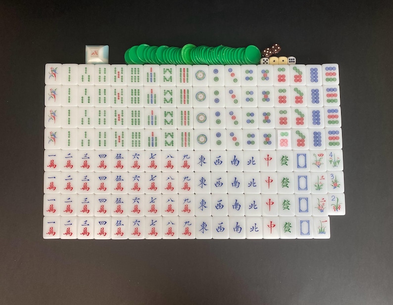 May include: A complete Mahjong set with white tiles featuring Chinese characters and symbols in red, green, and blue. Includes green chips, dice, and a white wind indicator. The set is arranged on a black surface.